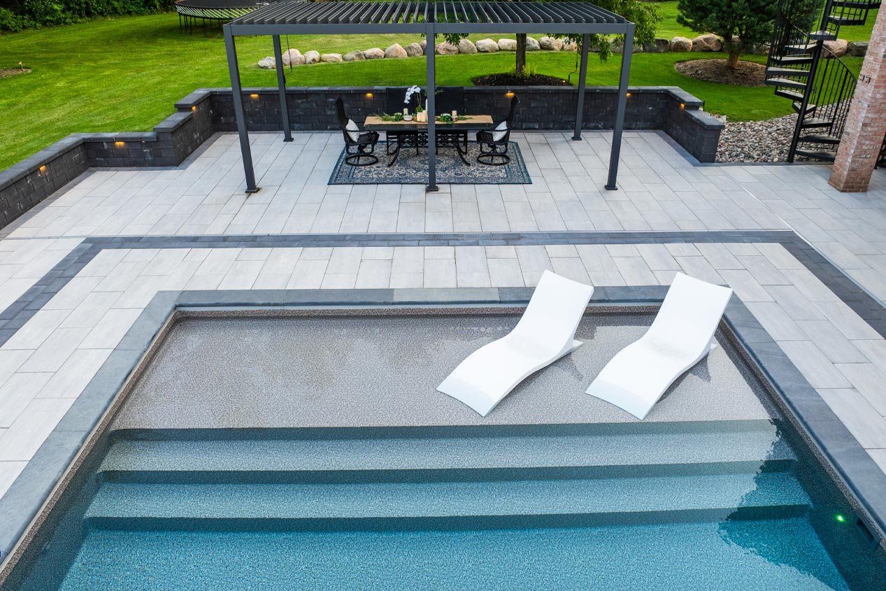 A backyard swimming pool with two white lounge chairs partially submerged in the shallow end, surrounded by a warm white and gray blend concrete paver pool deck with an accent band of charcoal-colored pavers arranged in a double sailor course.