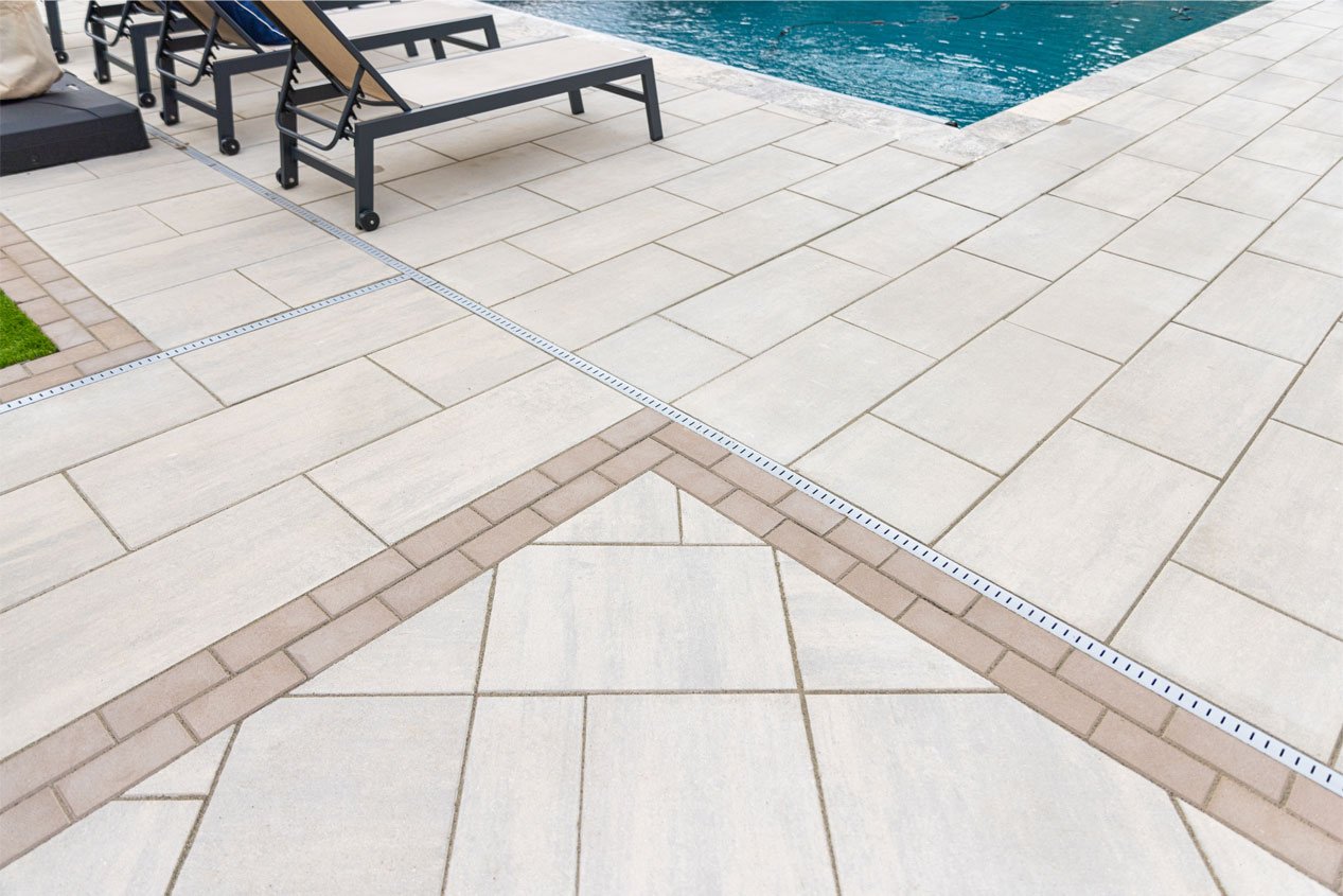 A warm white and beige blend concrete paver pool deck features pavers of many different sizes, with different sections laid at different angles and separated by a double sailor course border in a lightly contrasting tone.