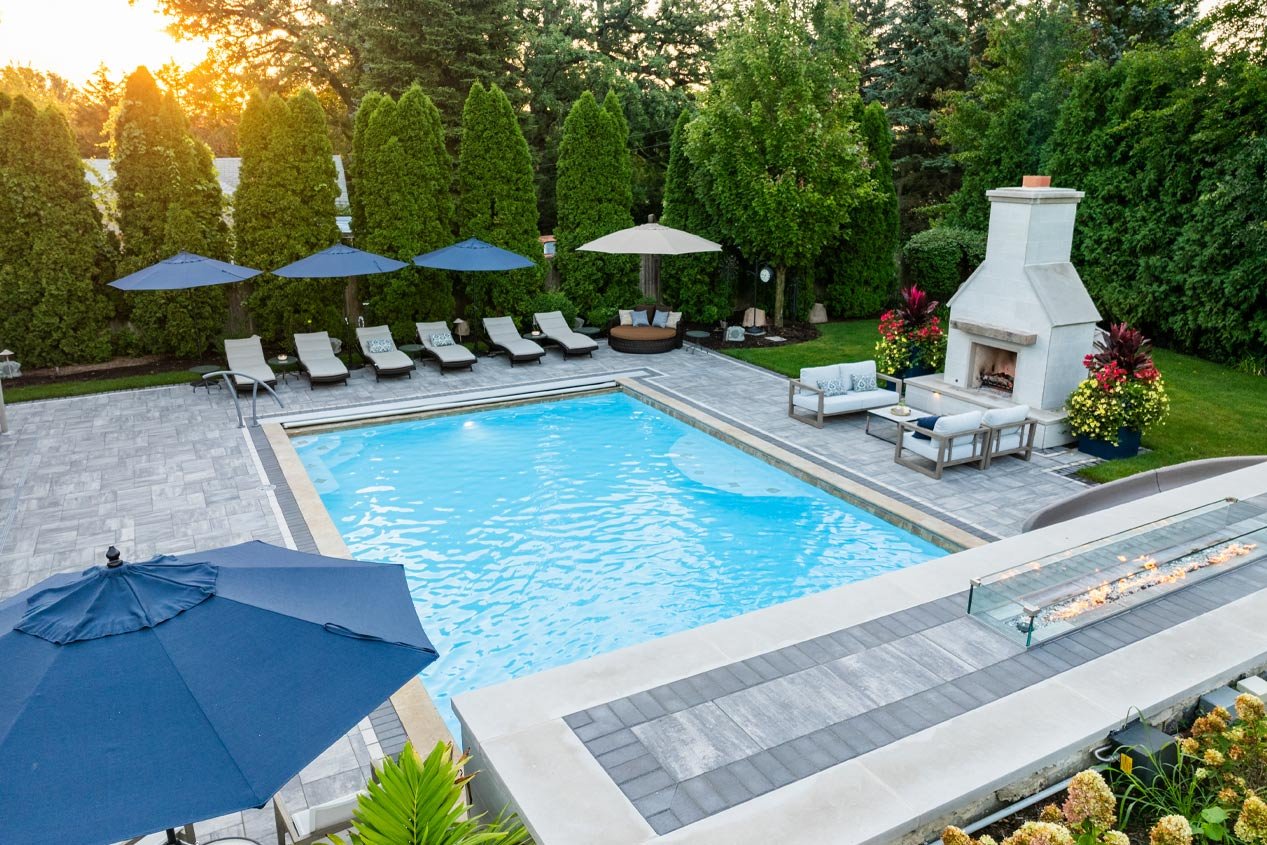 A rectangular pool surrounded by a concrete paver pool deck in gray variegated pavers has an impressive fire feature on the side that rises above the pool, with complementary concrete pavers on top of the top of the water feature that holds the gas fire feature.