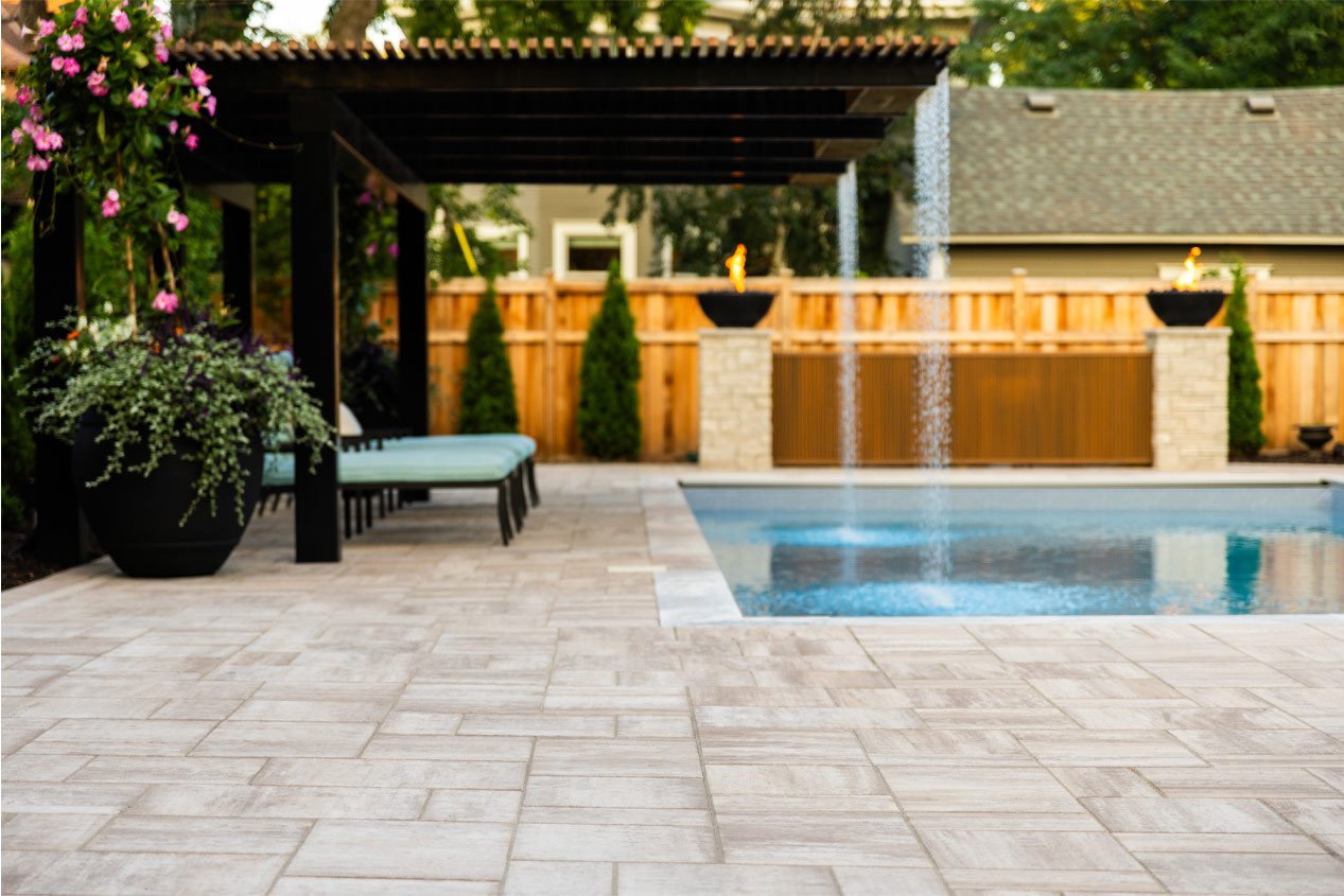 A light beige concrete paver pool deck includes a metal shade pergola with a built-in waterfall feature that falls into the pool.